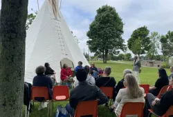 Group of people listening to Indigenous speakers, sitting in front of a tipi
