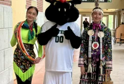 Two young women wearing Mi'kmaw regalia meet up with the UPEI mascot