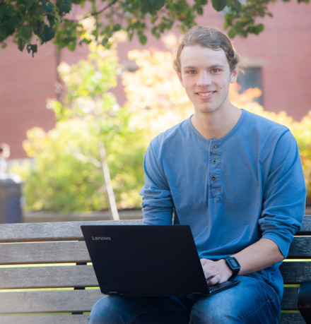 Jeremy sits on a bench outside with his laptop