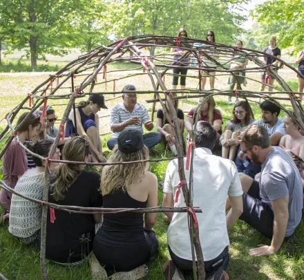 an indigenous elder and a group of people sitting inside the wooden frame of a sweat lodge