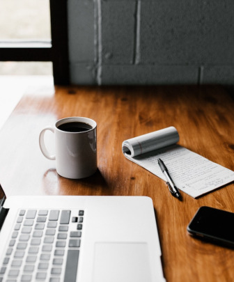 wooden table with a laptop computer, cup of coffee, cellular phone, and notepad