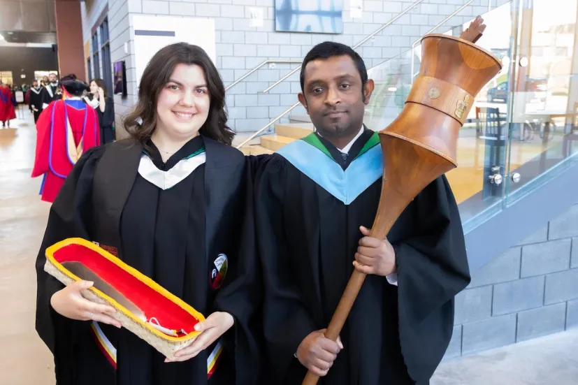 two people wearing black graduation gowns holding a mace and an eagle feather