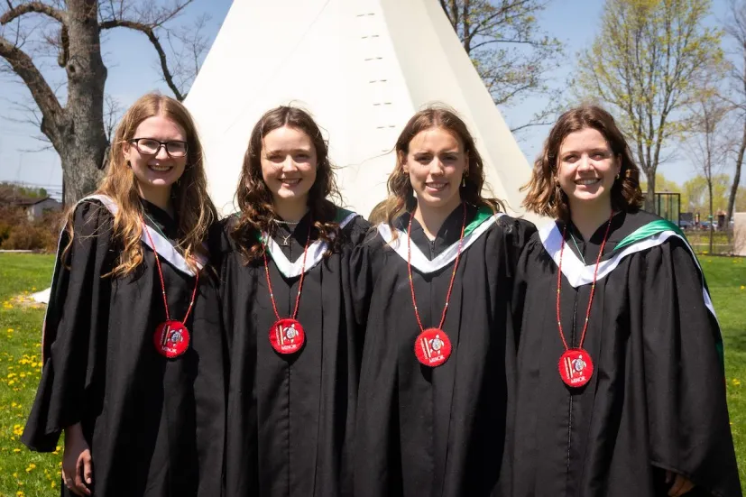 four graduates wearing black graduation gowns and red medallions