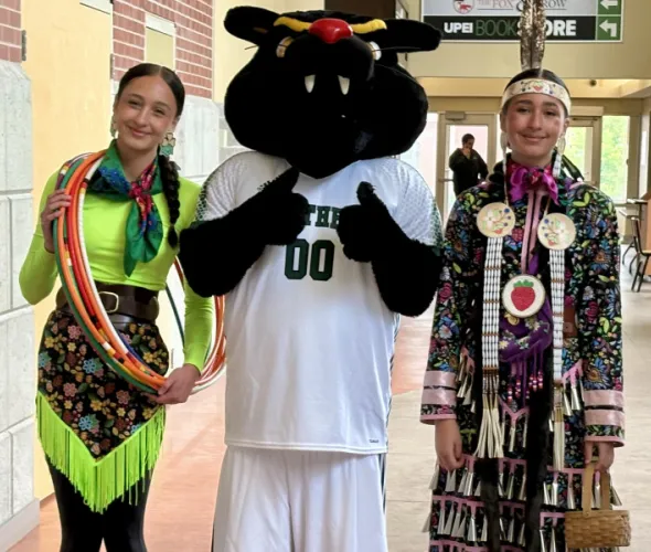 Two young women wearing Mi'kmaw regalia meet up with the UPEI mascot